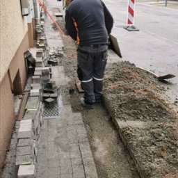 Rostalco Sp. z o.o. - Pracownik w kasku układa szarą kostkę brukową na chodniku przy budynku, widoczny wykop obok ułożonej części, narzędzia i pomarańczowy kabel w tle.