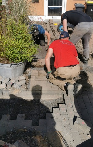 Trzech pracowników układa szarą kostkę brukową na podwórku przed domem; widoczny fragment ułożonej ścieżki, donica z rośliną i cień osoby robiącej zdjęcie.