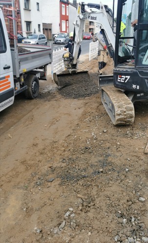 Dwa małe białe i czarne bagerki pracują na ulicy, jeden z pracownikiem w kasku, obok stoi samochód z przyczepą wypełnioną żwirem.