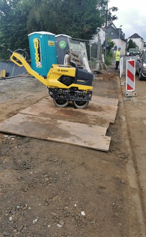Żółty walec drogowy BOMAG na placu budowy, stojący na drewnianych płytach, w tle przenośne toalety i pracownik w kamizelce odblaskowej.