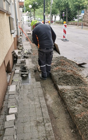 Pracownik w kasku układa szarą kostkę brukową na chodniku przy budynku, widoczny wykop obok ułożonej części, narzędzia i pomarańczowy kabel w tle.