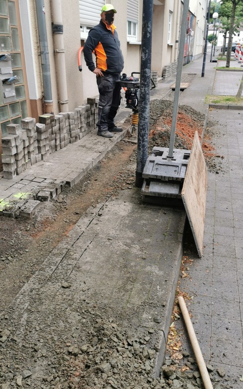 Pracownik z zagęszczarką skaczącą przygotowuje podłoże pod chodnik z kostki betonowej, stos kostki ułożony wzdłuż krawędzi, widoczne ślady po wykopie.