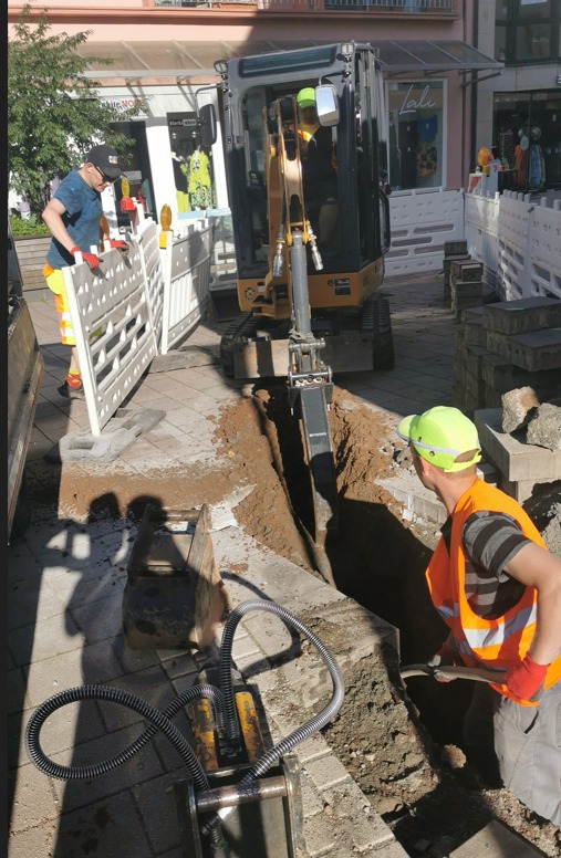 Mini koparka wykonuje wąski wykop w chodniku, pracownicy w odblaskowych kamizelkach zabezpieczają teren białymi barierkami w otoczeniu budynków handlowych.