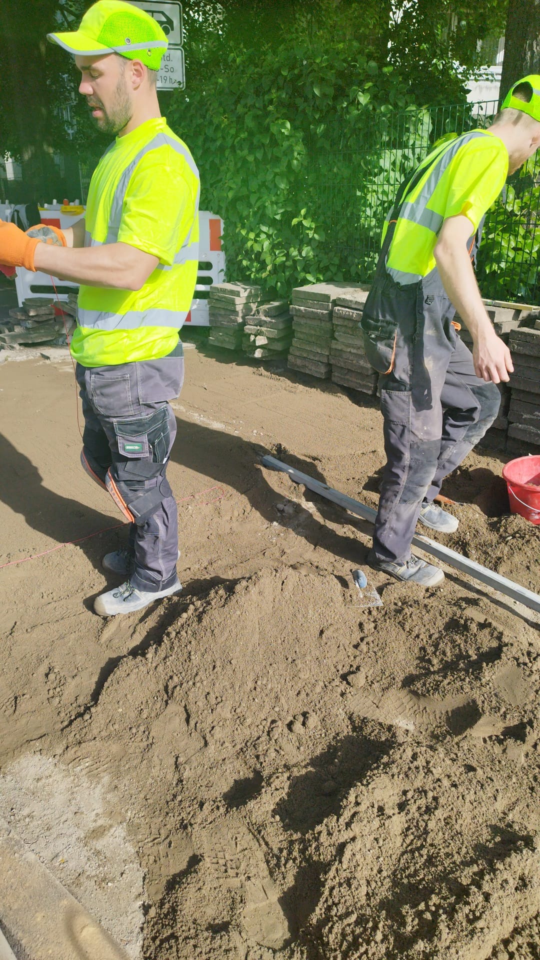 Dwóch robotników w odblaskowych ubraniach przygotowuje podłoże pod nawierzchnię z kostki, widoczne hałdy piasku, ułożone kostki i narzędzia.