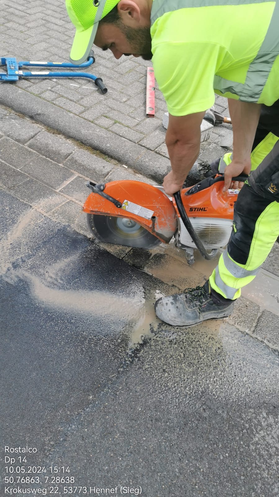 Pracownik w odblaskowej odzieży tnie piłą spalinową Stihl mokrą kostkę brukową, tworząc błoto z pyłu i wody.