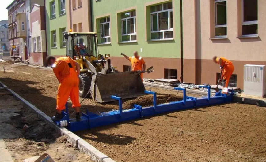 Ekipa budowlana w pomarańczowych kombinezonach wyrównuje podłoże pod chodnik przy użyciu niebieskiej listwy wibracyjnej, z żółtą ładowarką w tle i budynkami w tle.