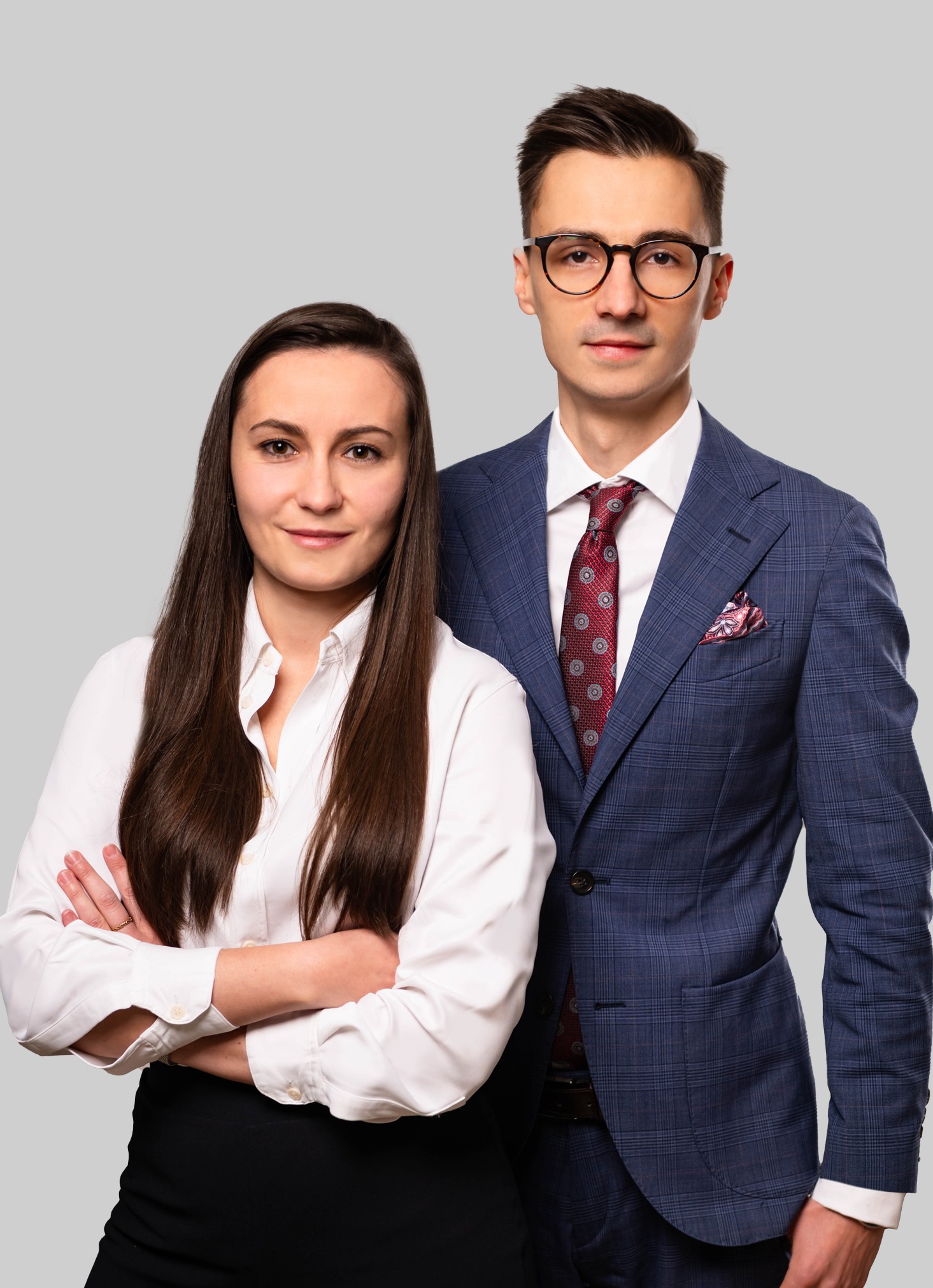 Portret dwójki prawników: kobieta z założonymi rękami w białej koszuli i mężczyzna w niebieskiej kraciastej marynarce z bordowym krawatem na szarym tle.