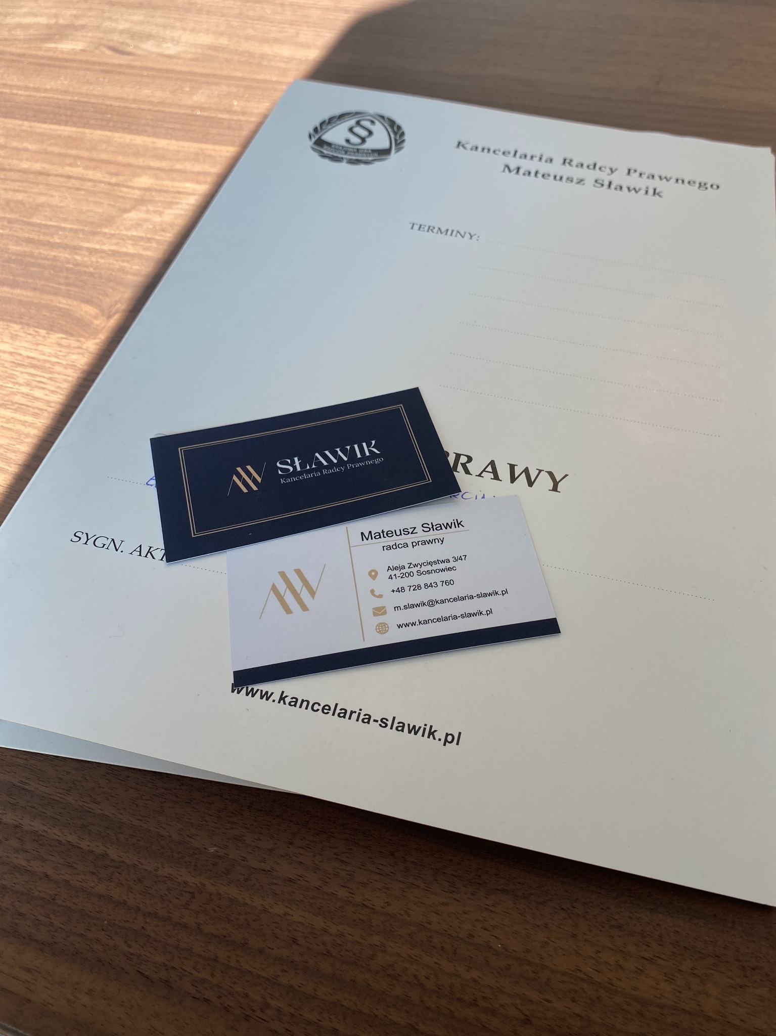Dokumenty Kancelarii Radcy Prawnego Mateusza Sławika: firmowy segregator, wizytówki na drewnianym biurku. Widoczne logo i dane kontaktowe.