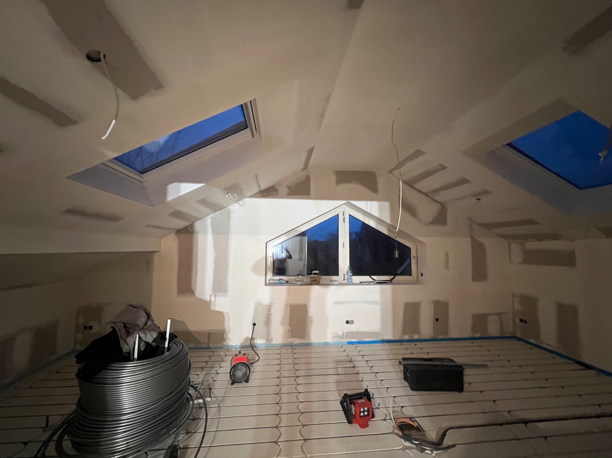 Poddasze w trakcie prac wykończeniowych: szpachlowane ściany, okna dachowe i rozłożona instalacja ogrzewania podłogowego z widocznymi narzędziami.