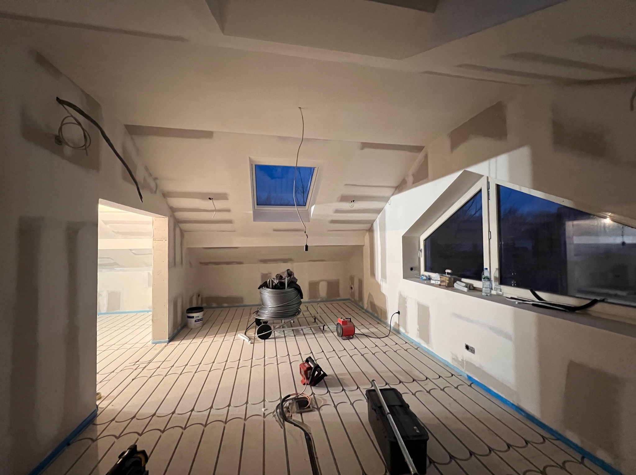 Poddasze w trakcie wykańczania: szpachlowane ściany, okno dachowe, instalacja ogrzewania podłogowego z rozłożonymi rurami, narzędzia i materiały budowlane.