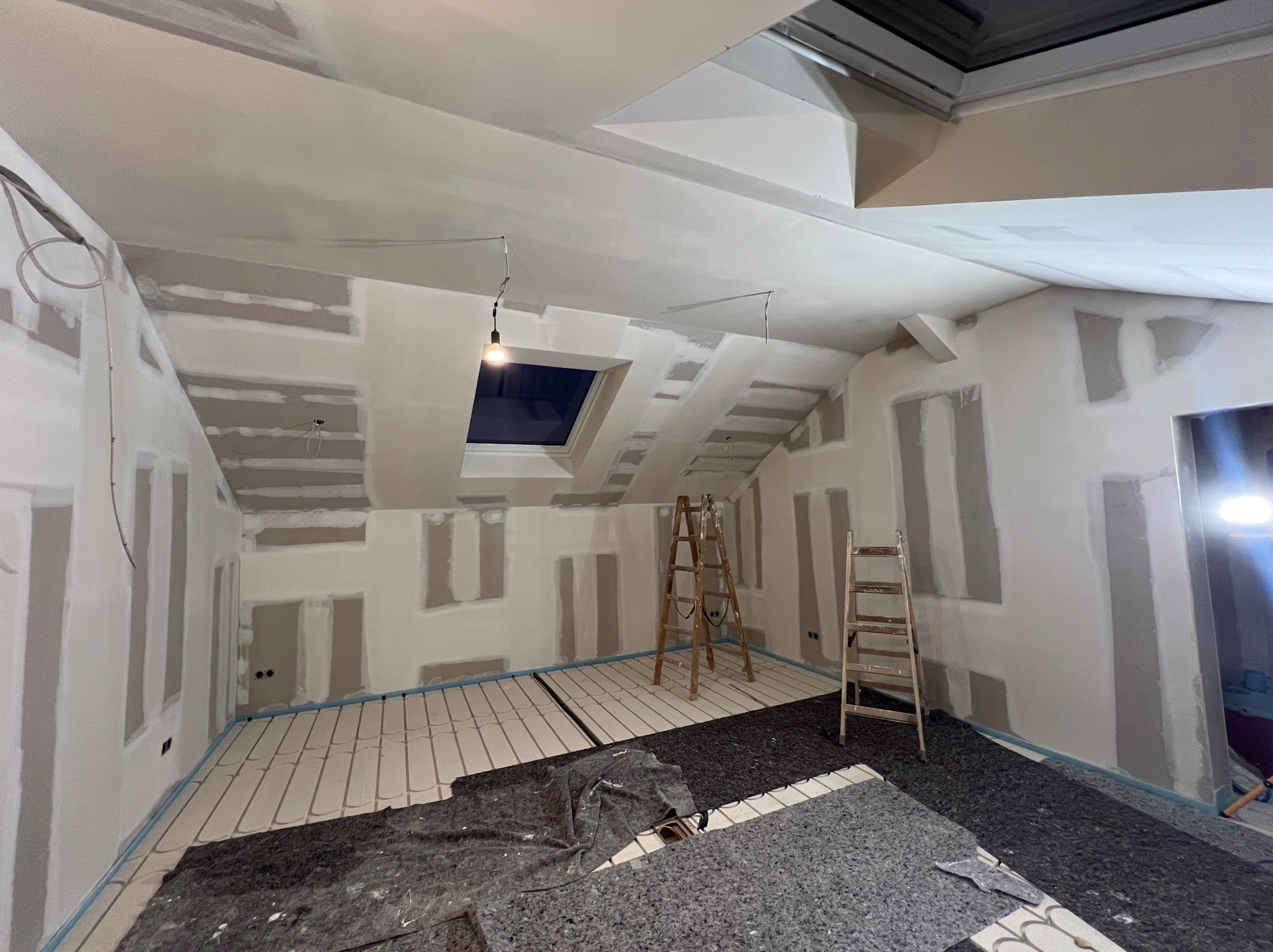 Poddasze w trakcie prac wykończeniowych: ściany z fragmentami płyt gips-karton, instalacja ogrzewania podłogowego, drabiny. Widok na okno dachowe i surowe oświetlenie.