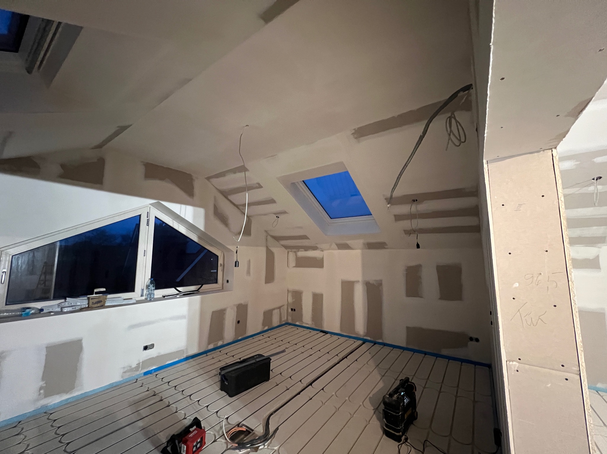 Poddasze w trakcie prac wykończeniowych: ściany częściowo wyszpachlowane, widoczne rury ogrzewania podłogowego, okno dachowe i okno w ścianie szczytowej.