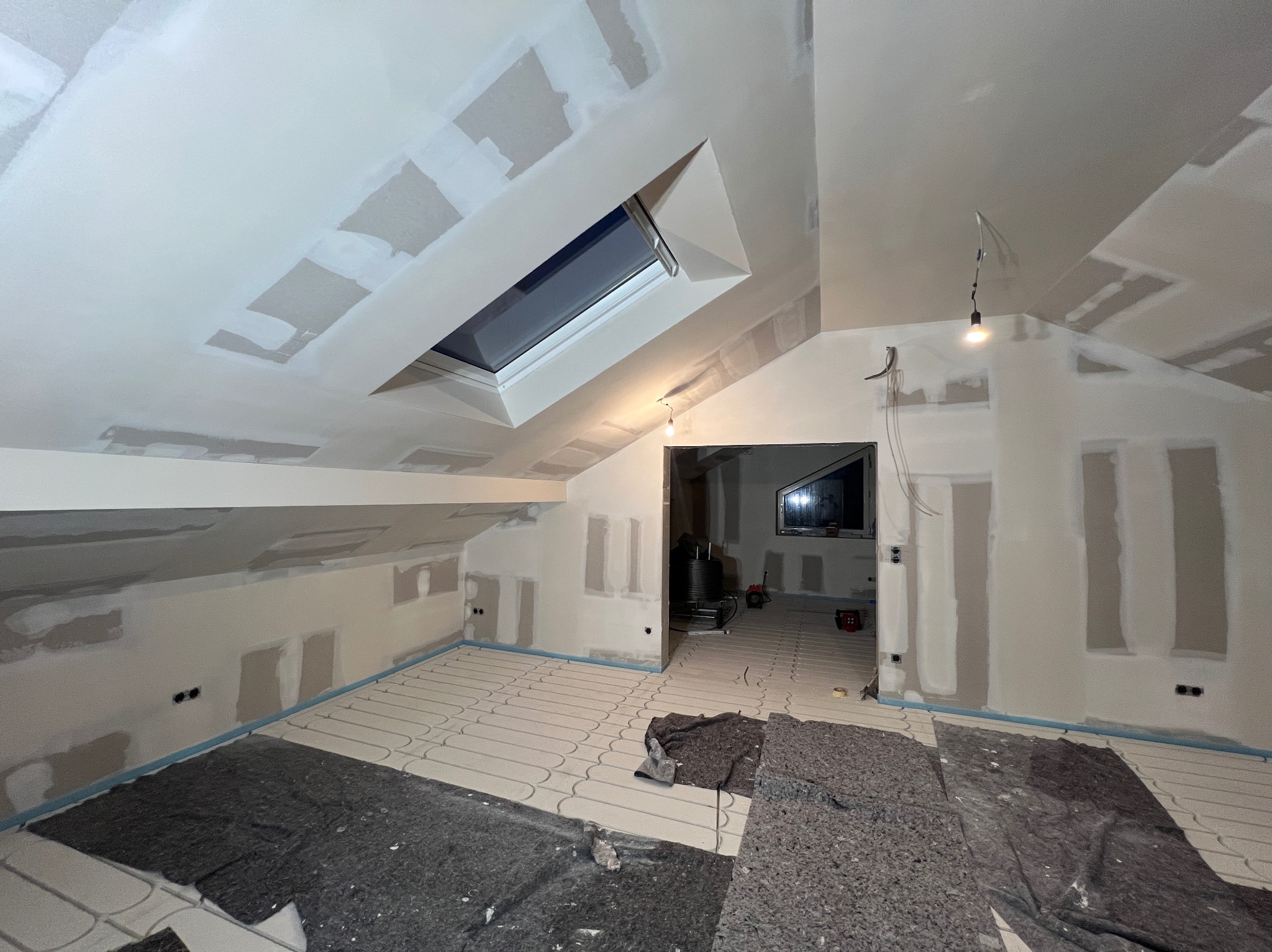 Poddasze w trakcie prac wykończeniowych: szpachlowane ściany, okno dachowe, instalacja ogrzewania podłogowego, widoczne kable i gniazdka elektryczne. Etap przygotowania do malowania.