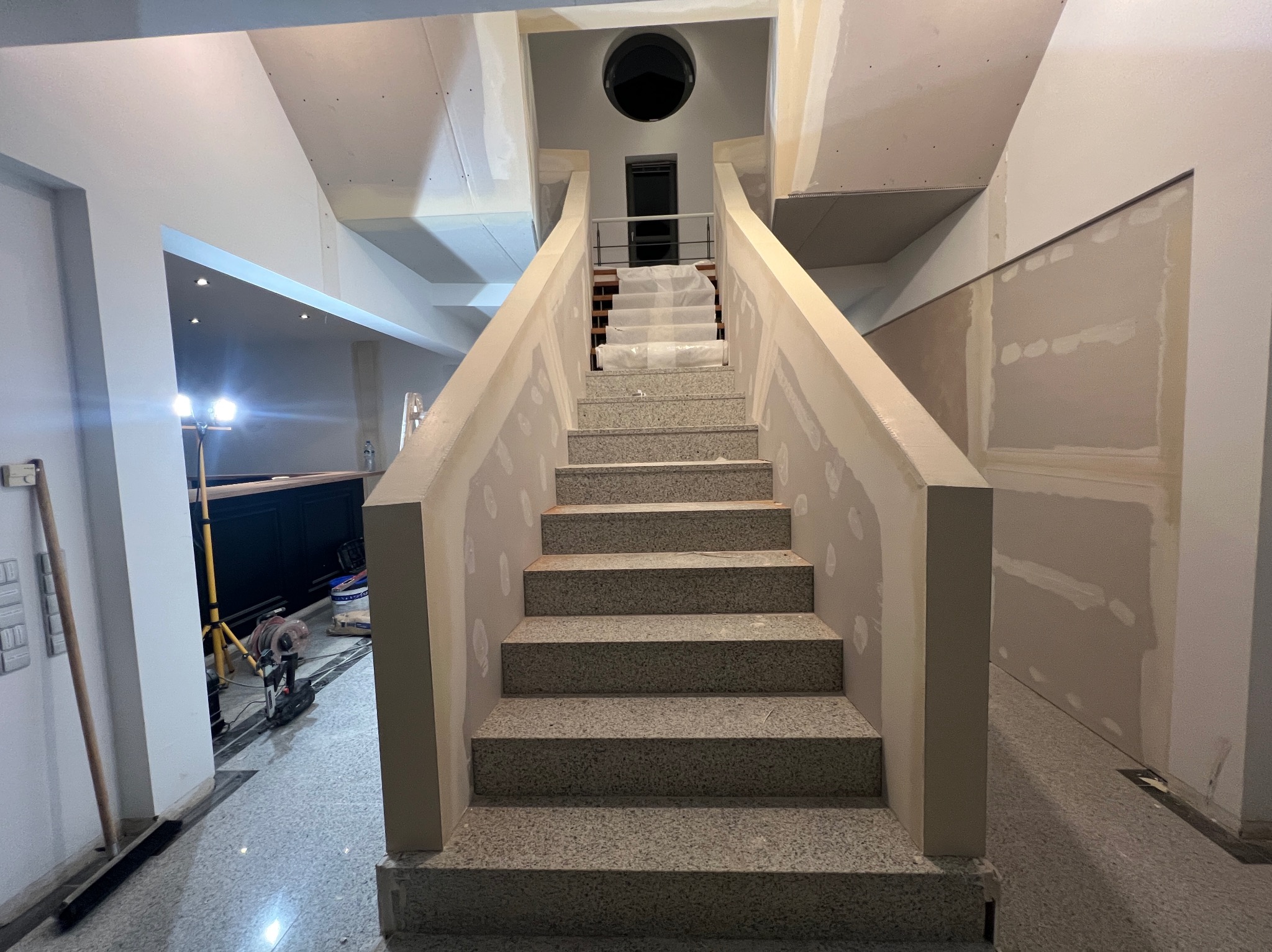Klatka schodowa w trakcie prac wykończeniowych: szpachlowane ściany, zabezpieczone schody, widoczne narzędzia. Wnętrze w jasnych barwach, surowy stan.