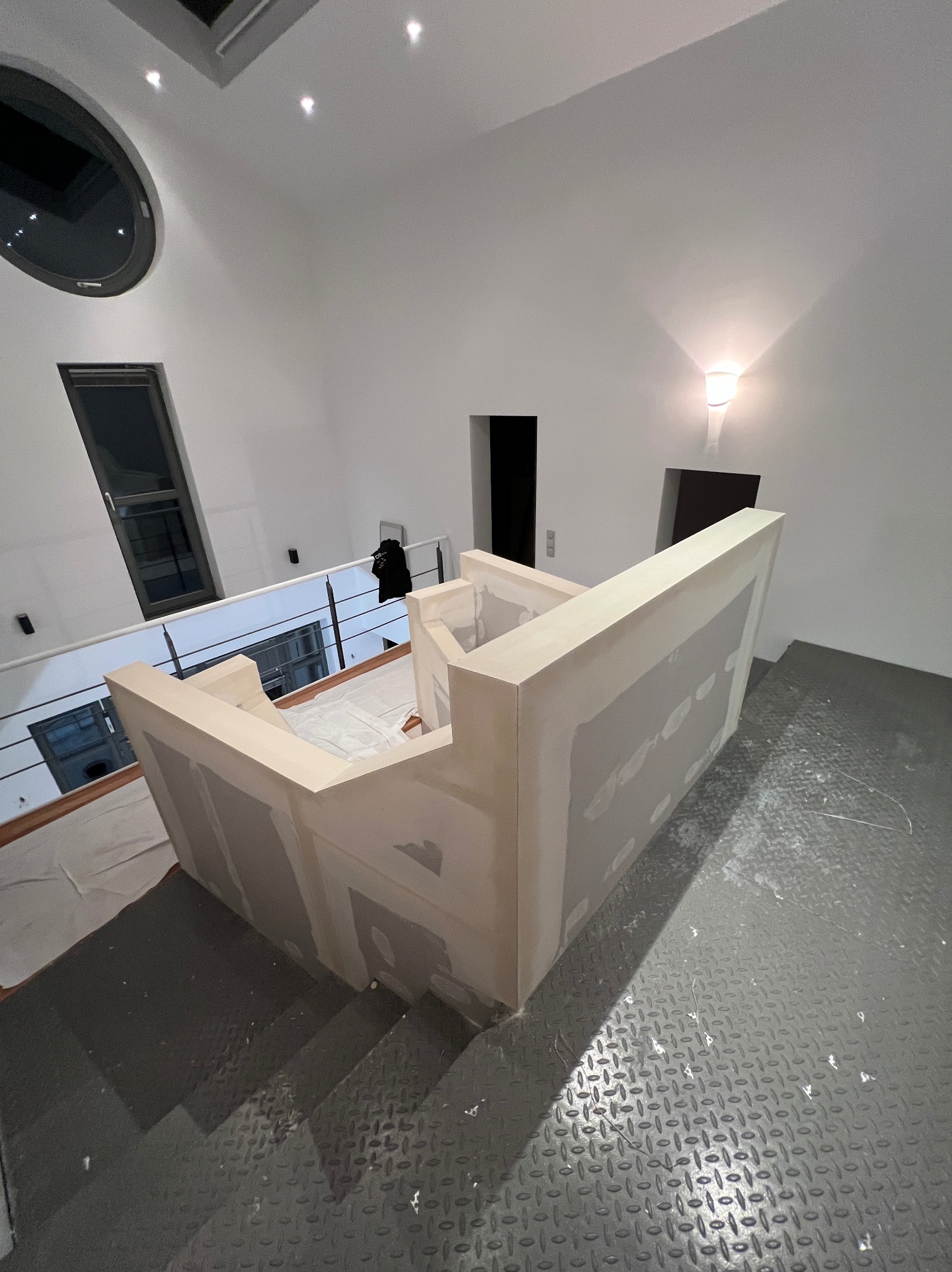 Wnętrze w trakcie wykończenia: zabudowa schodów z płyt karton-gips, widok z góry na metalowe schody, białe ściany, okno owalne i lampa ścienna.