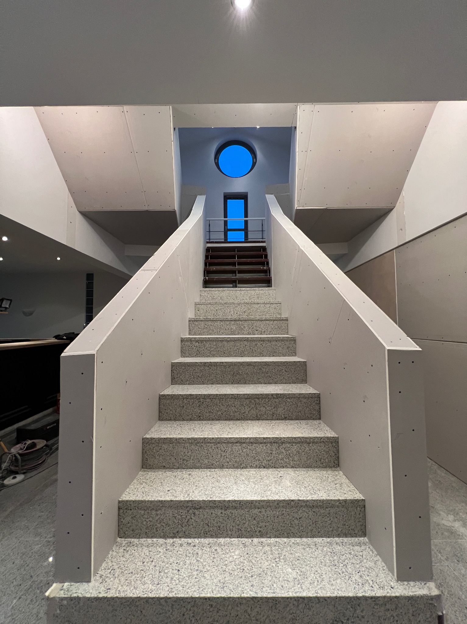 Klatka schodowa w trakcie montażu płyt kartonowo-gipsowych, widoczne szare płyty i schody z lastryko, okrągłe okno w tle.