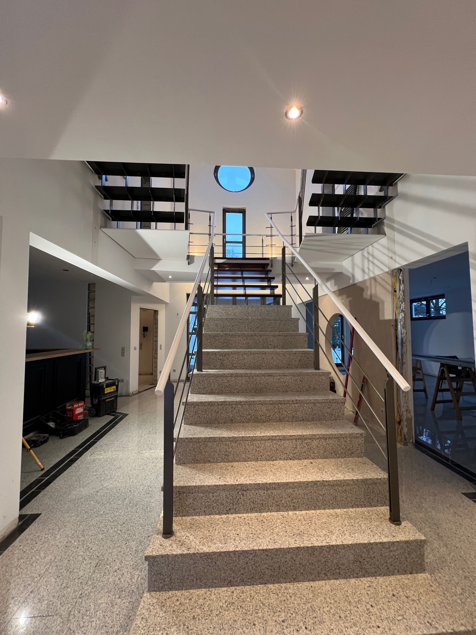 Wnętrze domu w trakcie wykończenia: schody z metalową balustradą, widoczne narzędzia i materiały budowlane, okrągłe okno w suficie. Prace wykończeniowe w toku.