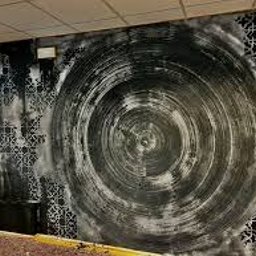 Samsobieremontrobie - Ściana z abstrakcyjnym wzorem w odcieniach szarości i czerni, przypominającym wir, z fragmentami dekoracyjnego szablonu w narożnikach, widoczne elementy instalacji elektrycznej i fragment podłogi.