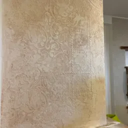 Beżowa ściana z dekoracyjną, wypukłą fakturą w stylu vintage, zabezpieczona folią malarską u dołu.