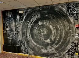 Ściana z abstrakcyjnym wzorem w odcieniach szarości i czerni, przypominającym wir, z fragmentami dekoracyjnego szablonu w narożnikach, widoczne elementy instalacji elektrycznej i fragment podłogi.