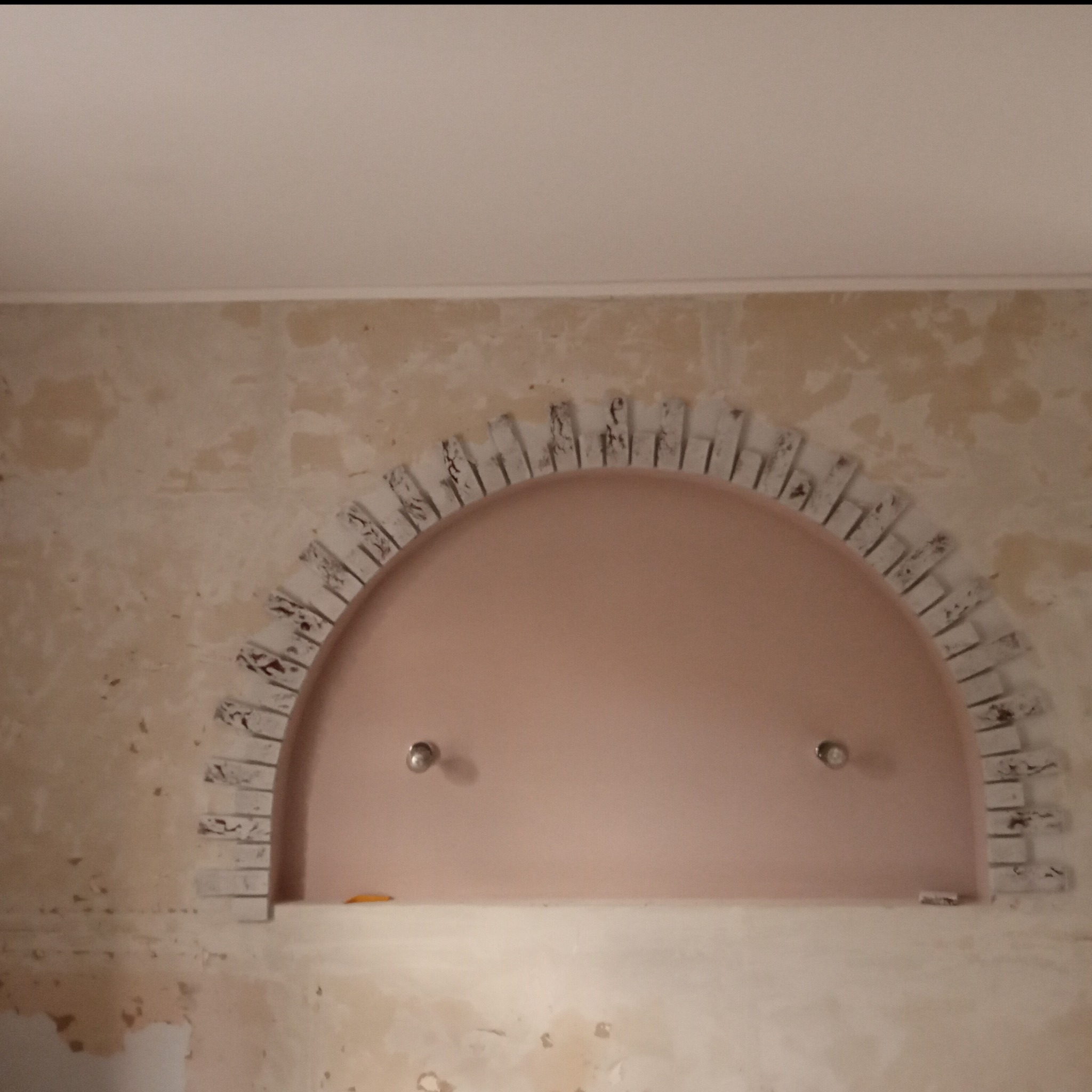 Nieregularny łuk z cegiełek nad wnęką w ścianie, w tle ściana w trakcie przygotowywania do malowania, widoczne dwa punkty świetlne.