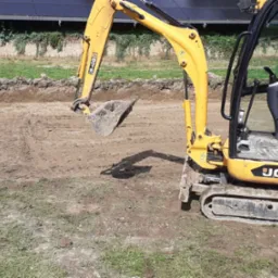 Żółta mini koparka JCB przygotowuje wykop pod fundamenty, widoczne ślady gąsienic w ziemi i wykopany obszar z równymi ścianami, w tle zieleń i panele słoneczne.