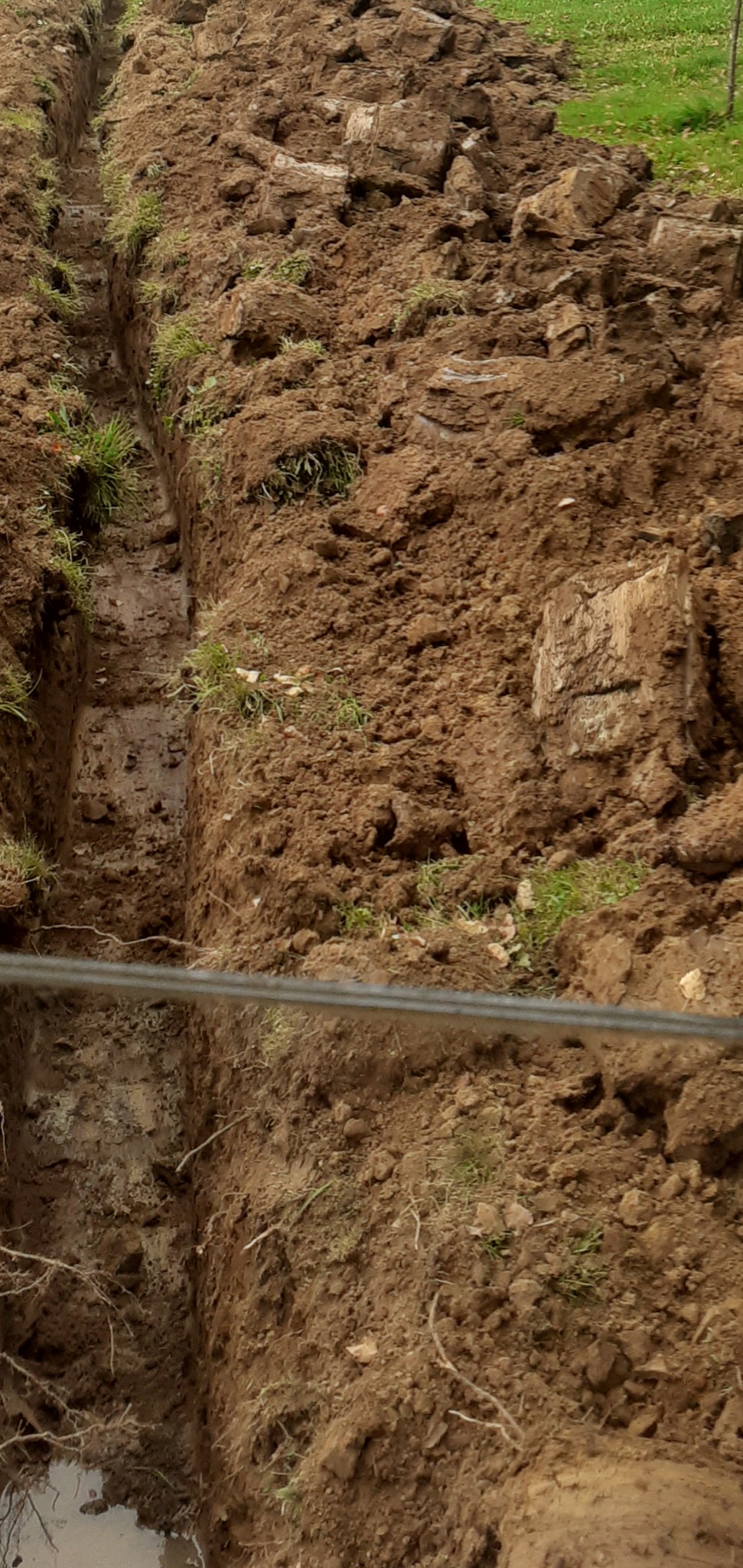Wykopany wąski rów w ziemi, widoczne ślady wody na dnie, obok pryzma wykopanej ziemi z resztkami trawy, w kadrze widoczny przewód.