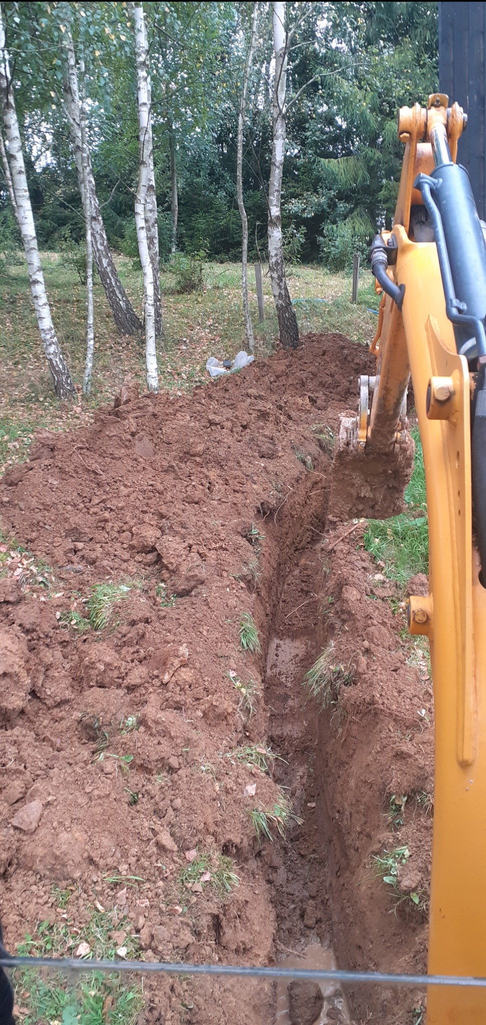 Wykopany rów w Ustrzykach Dolnych, widoczny fragment koparki i drzewa w tle, ziemia brązowa, na dnie rowu woda.