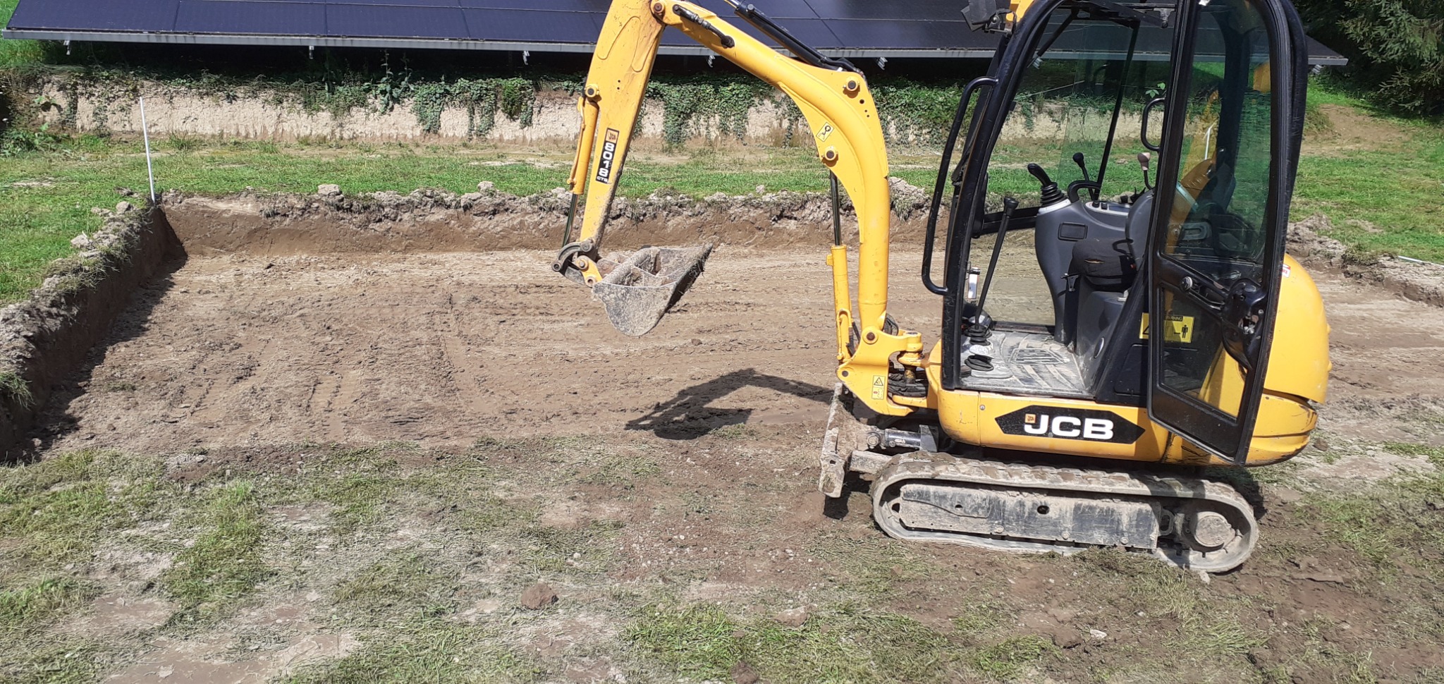 Żółta mini koparka JCB przygotowuje wykop pod fundamenty, widoczne ślady gąsienic w ziemi i wykopany obszar z równymi ścianami, w tle zieleń i panele słoneczne.