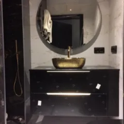 Nowoczesna łazienka z czarną szafką pod umywalkę, złotą, młotkowaną umywalką nablatową i okrągłym lustrem z podświetleniem LED.