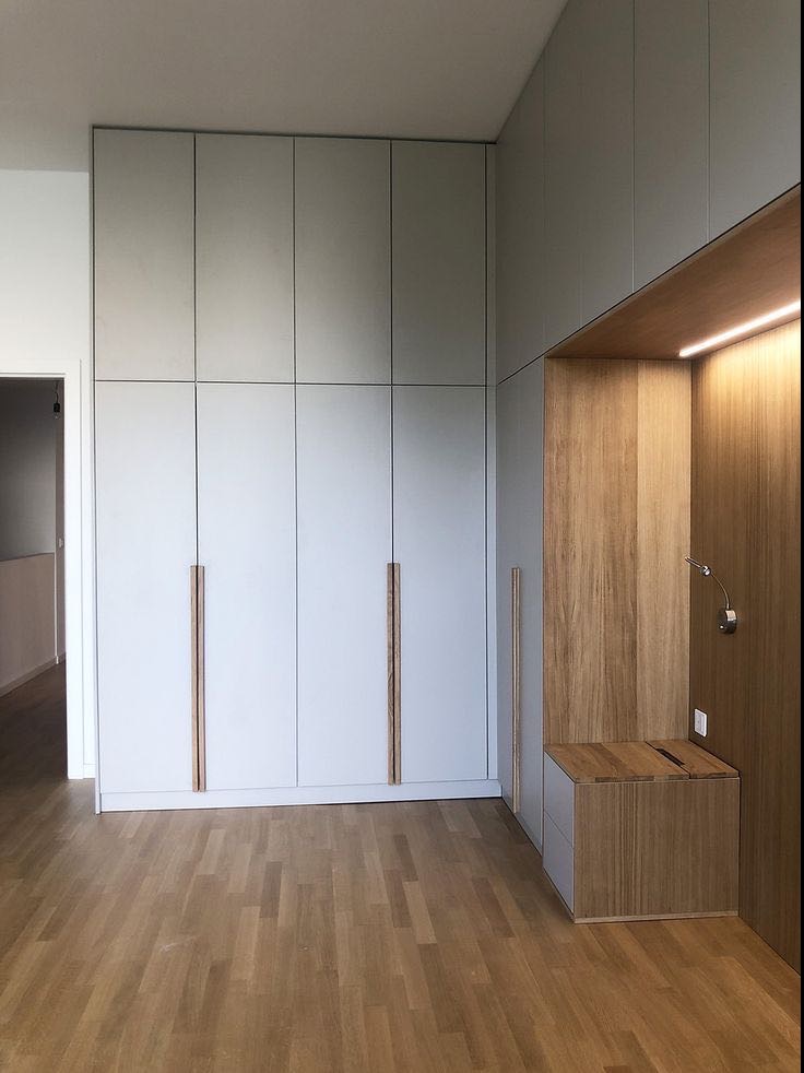 Wysoka, zabudowana szafa wnękowa z białymi frontami i długimi, drewnianymi uchwytami, połączona z drewnianą zabudową z oświetleniem LED i małą szafką nocną, na podłodze parkiet.