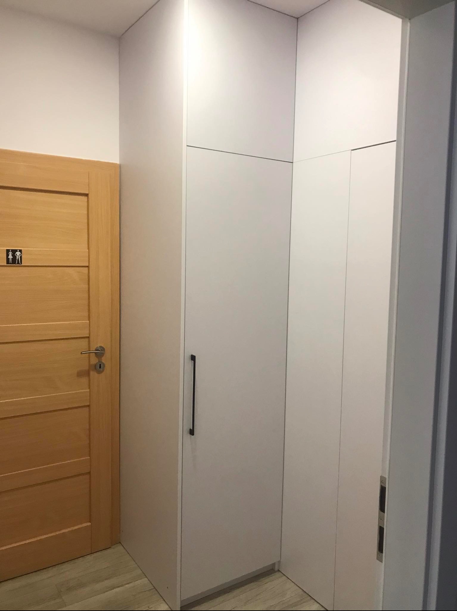 Biała szafa wnękowa z minimalistycznym uchwytem, sąsiadująca z drewnianymi drzwiami oznaczonymi piktogramem toalety, na tle jasnej podłogi.
