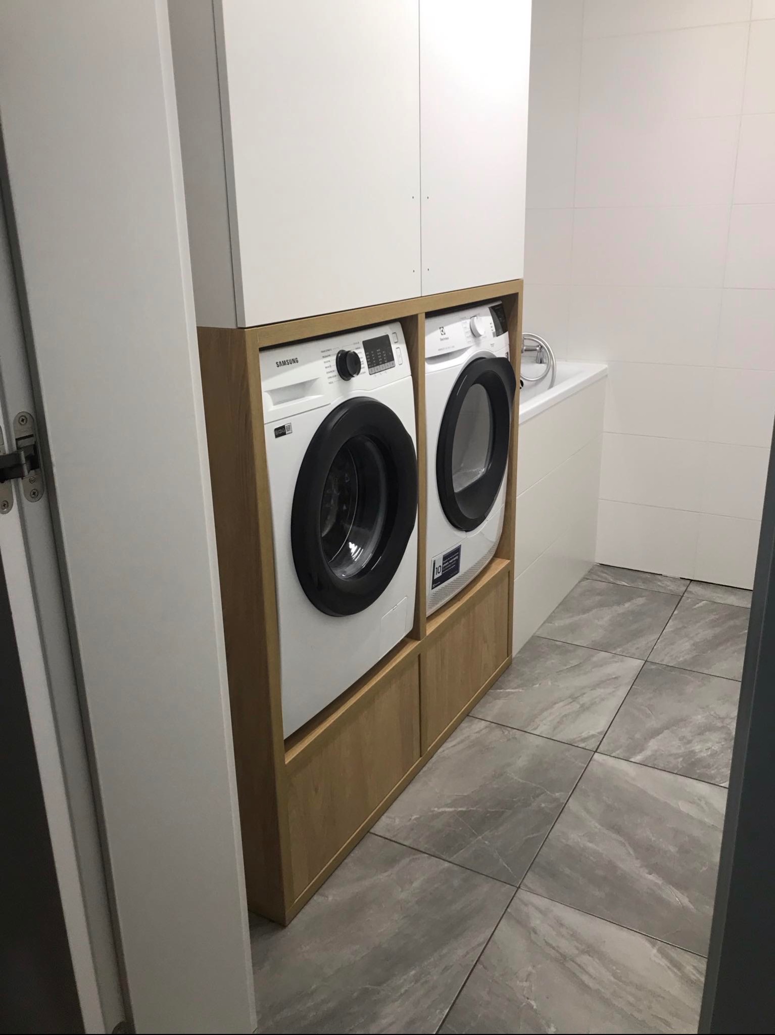 Dwie pralki (Samsung i Electrolux) zabudowane w drewnianej konstrukcji z białymi szafkami wiszącymi w łazience z szarą podłogą z płytek imitujących marmur.