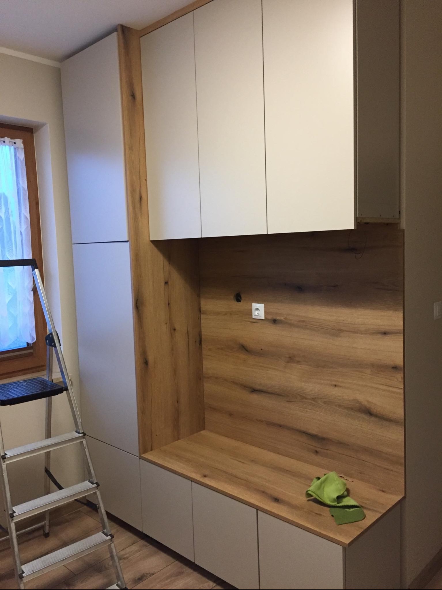 Wnęka z drewnianym panelem i gniazdkiem elektrycznym, połączona z szafkami w kolorze złamanej bieli, drabina malarska oparta o ścianę, zielona ściereczka leży na blacie.