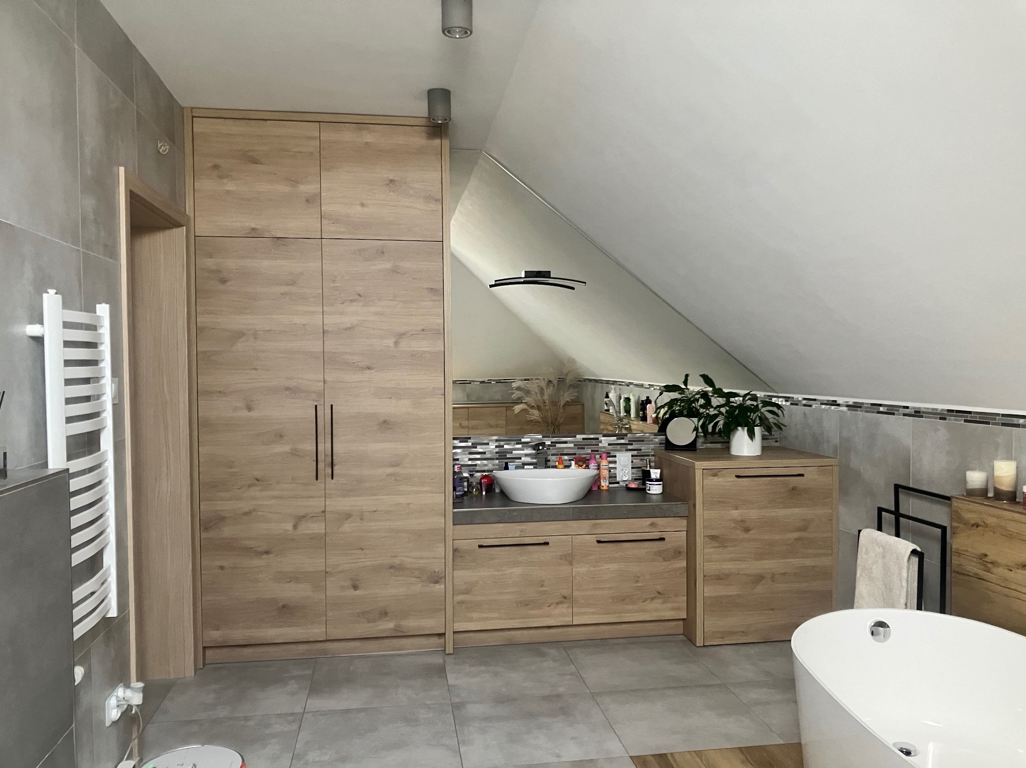 Jasna łazienka na poddaszu z drewnianą zabudową szaf, umywalką nablatową i wanną wolnostojącą.