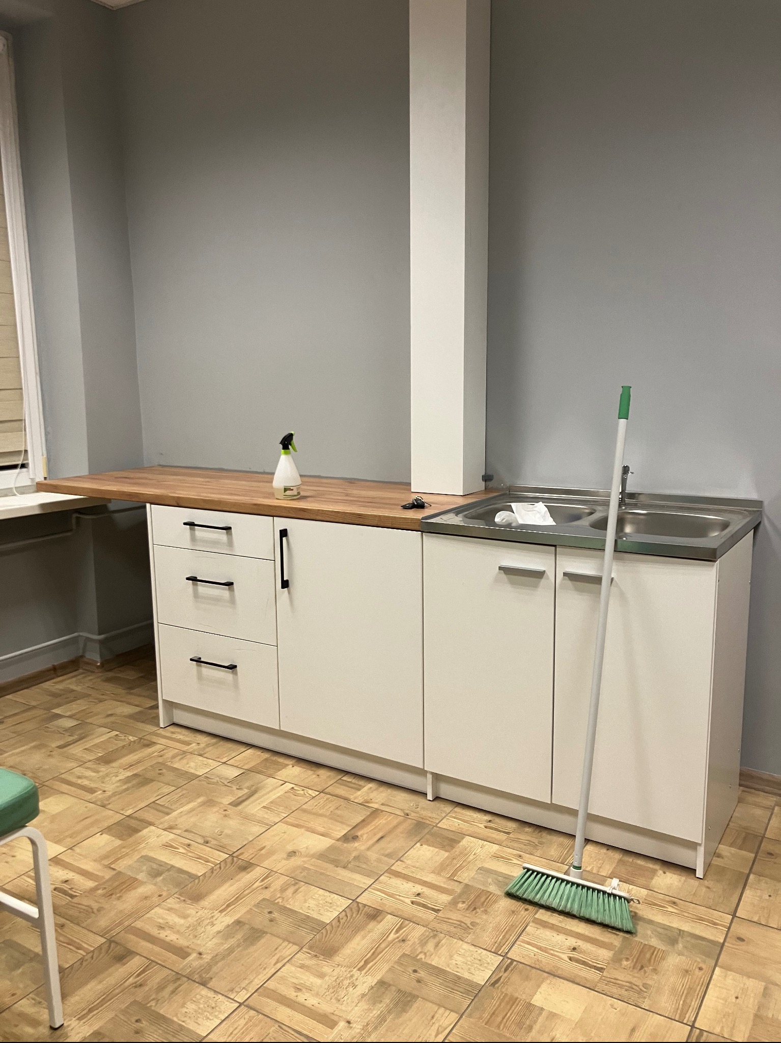 Prosta zabudowa kuchenna z drewnianym blatem i białymi frontami, uzupełniona dwukomorowym zlewem ze stali nierdzewnej, z widoczną butelką ze spryskiwaczem i szczotką do zamiatania na podłodze.