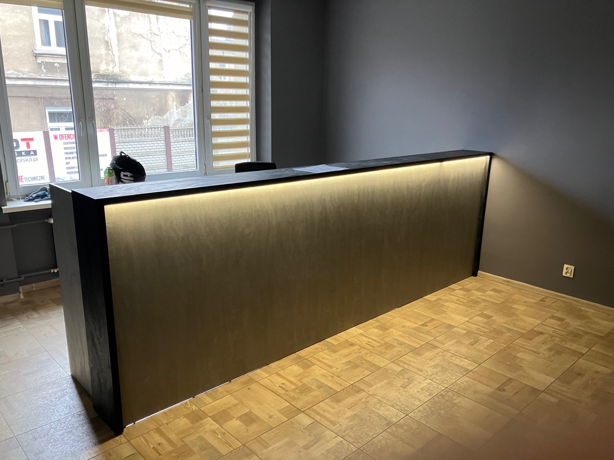 Recepcja z podświetlanym blatem w nowoczesnym biurze, widok z przodu, na blacie leżą dokumenty, w tle okno z roletami.