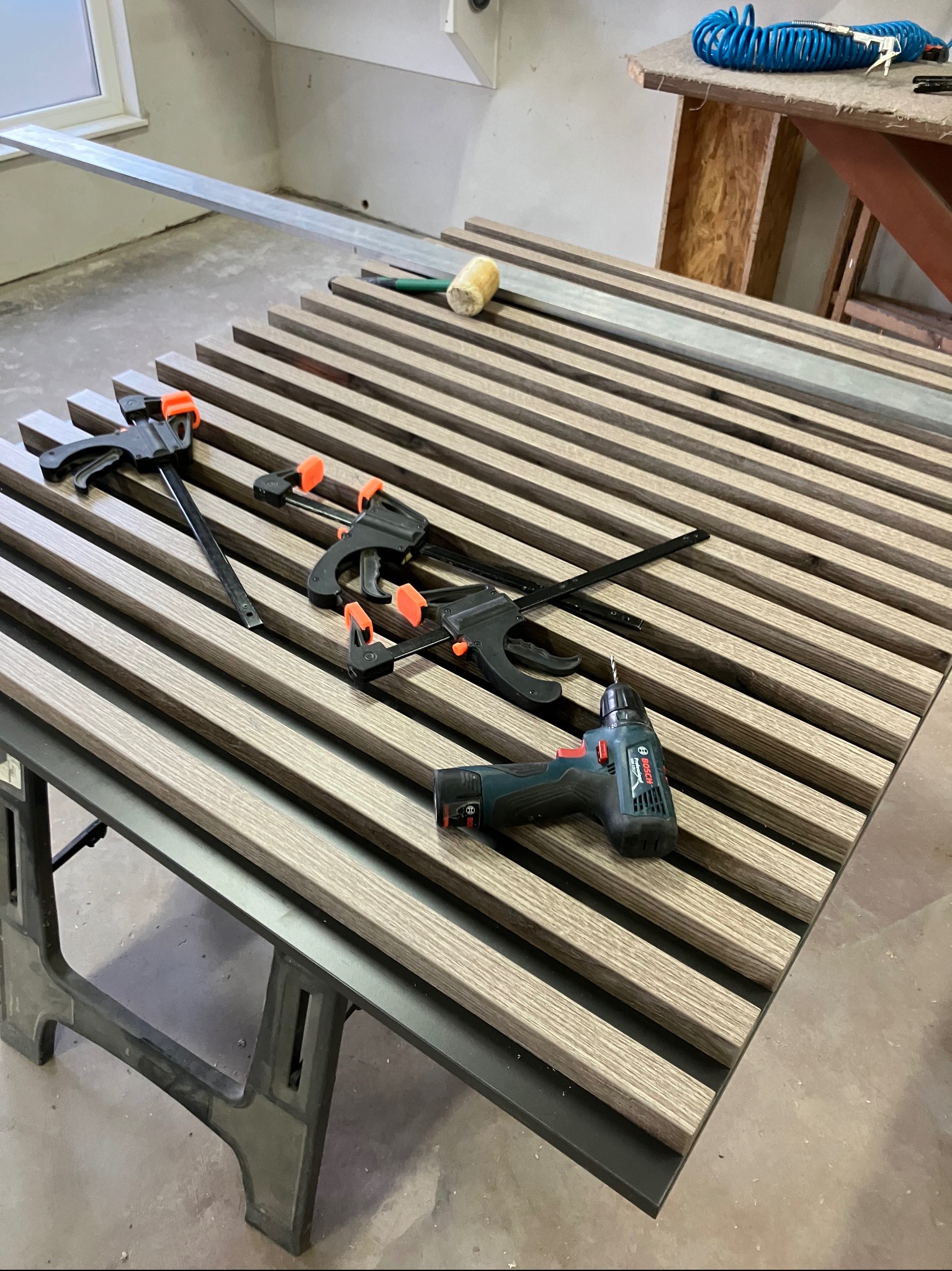 Panel z drewnianych listew, przytwierdzonych klamrami do ramy, na stole warsztatowym, w tle wiertarka akumulatorowa i narzędzia.