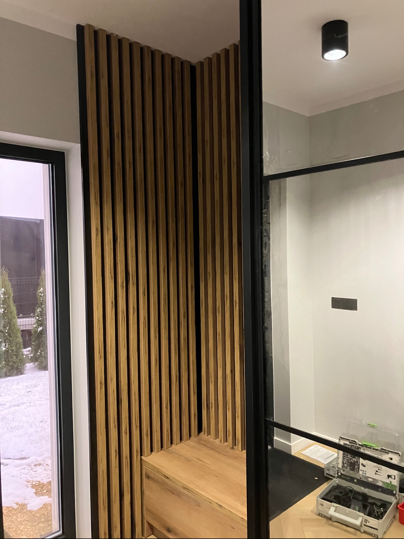 Drewniana ścianka działowa z pionowych lameli w czarnej ramie, z siedziskiem i szufladą, widok zza szklanej ściany, skrzynki narzędziowe w tle.