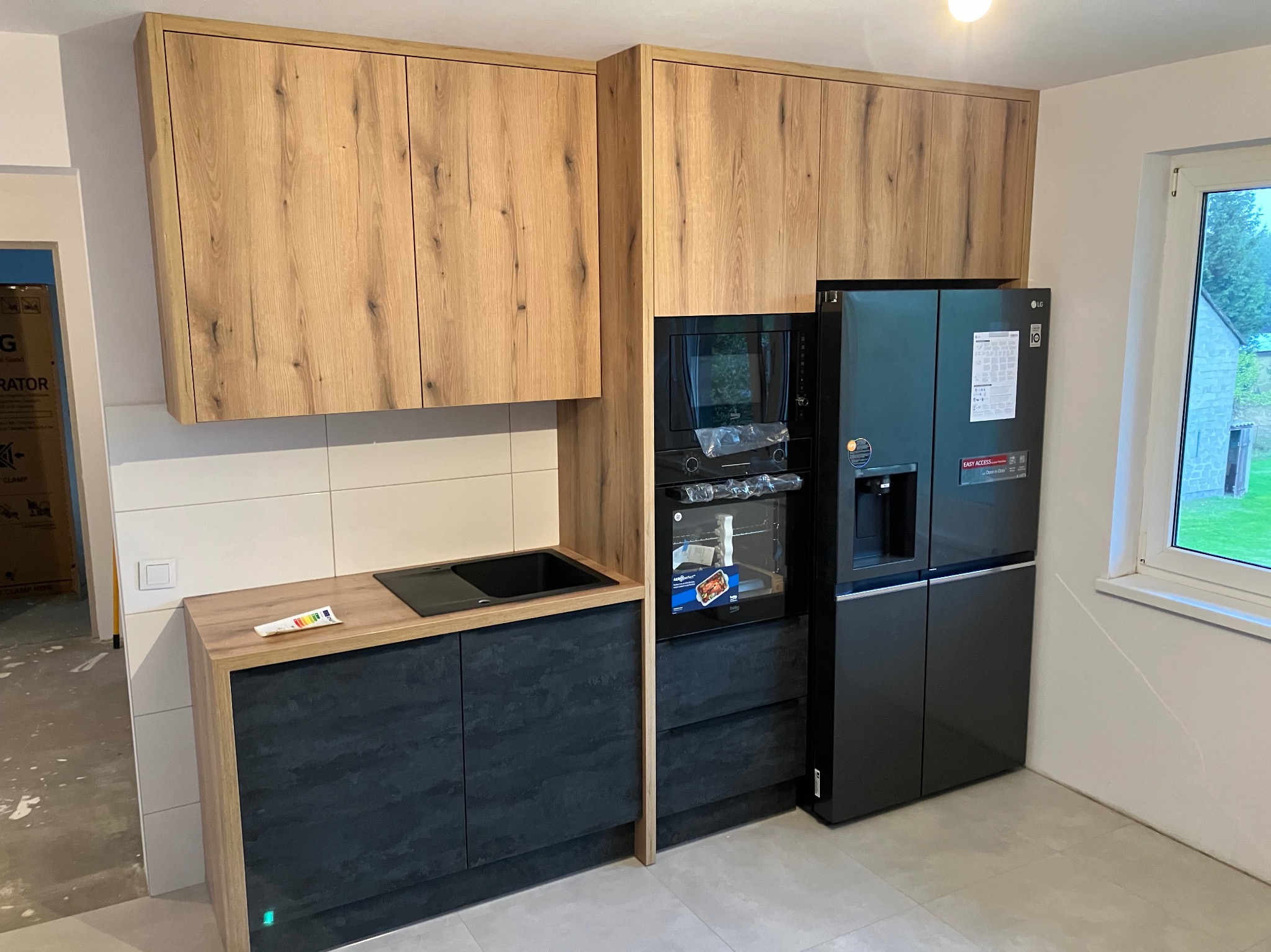 Fragment nowej kuchni z drewnianymi szafkami górnymi i czarnymi szafkami dolnymi, czarny zlew, piekarnik i duża lodówka LG obok okna.