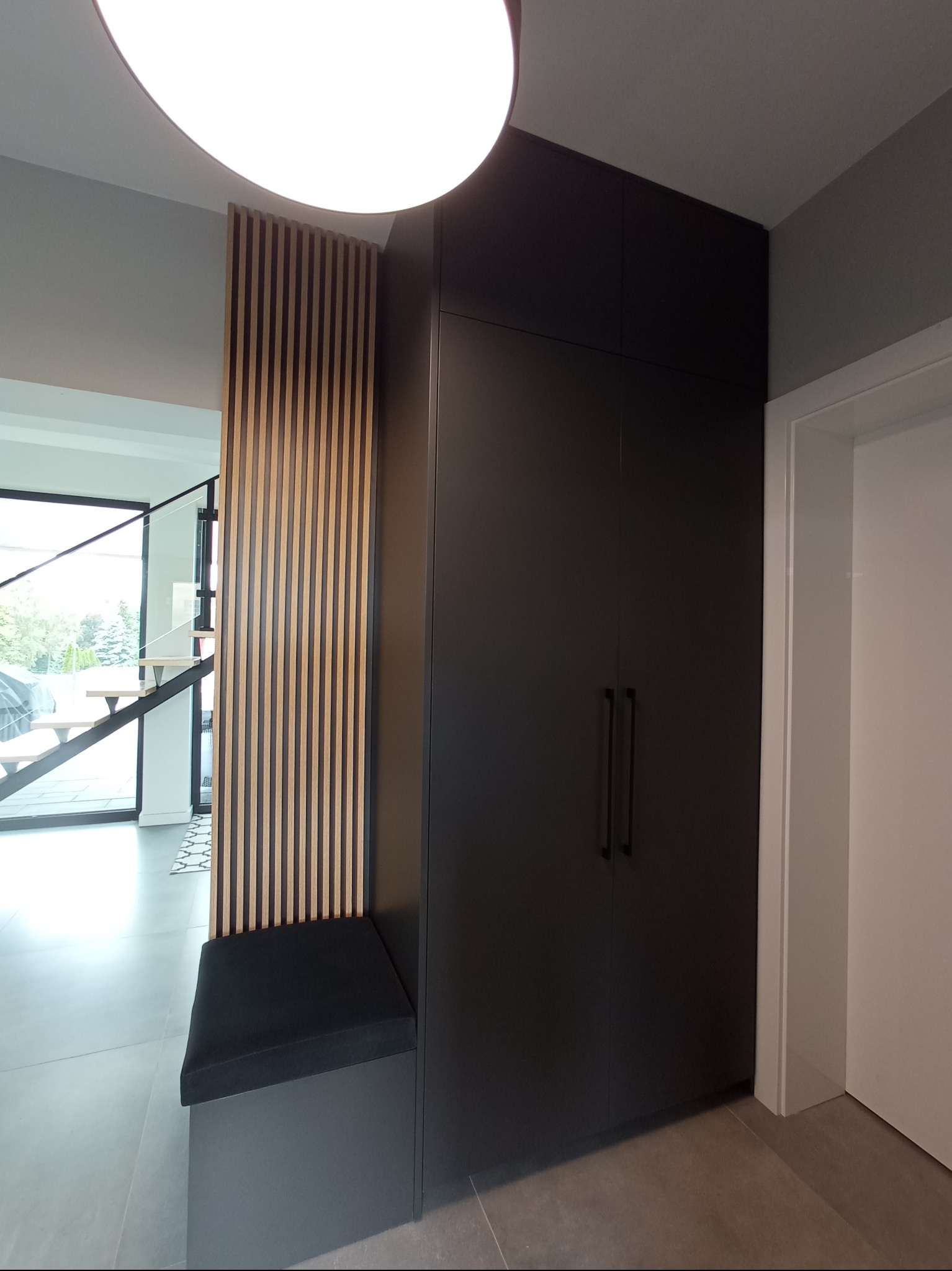 Ciemna szafa z długimi uchwytami, drewniane lamele na ścianie oraz siedzisko z czarną poduszką w nowoczesnym przedpokoju z widocznymi schodami.