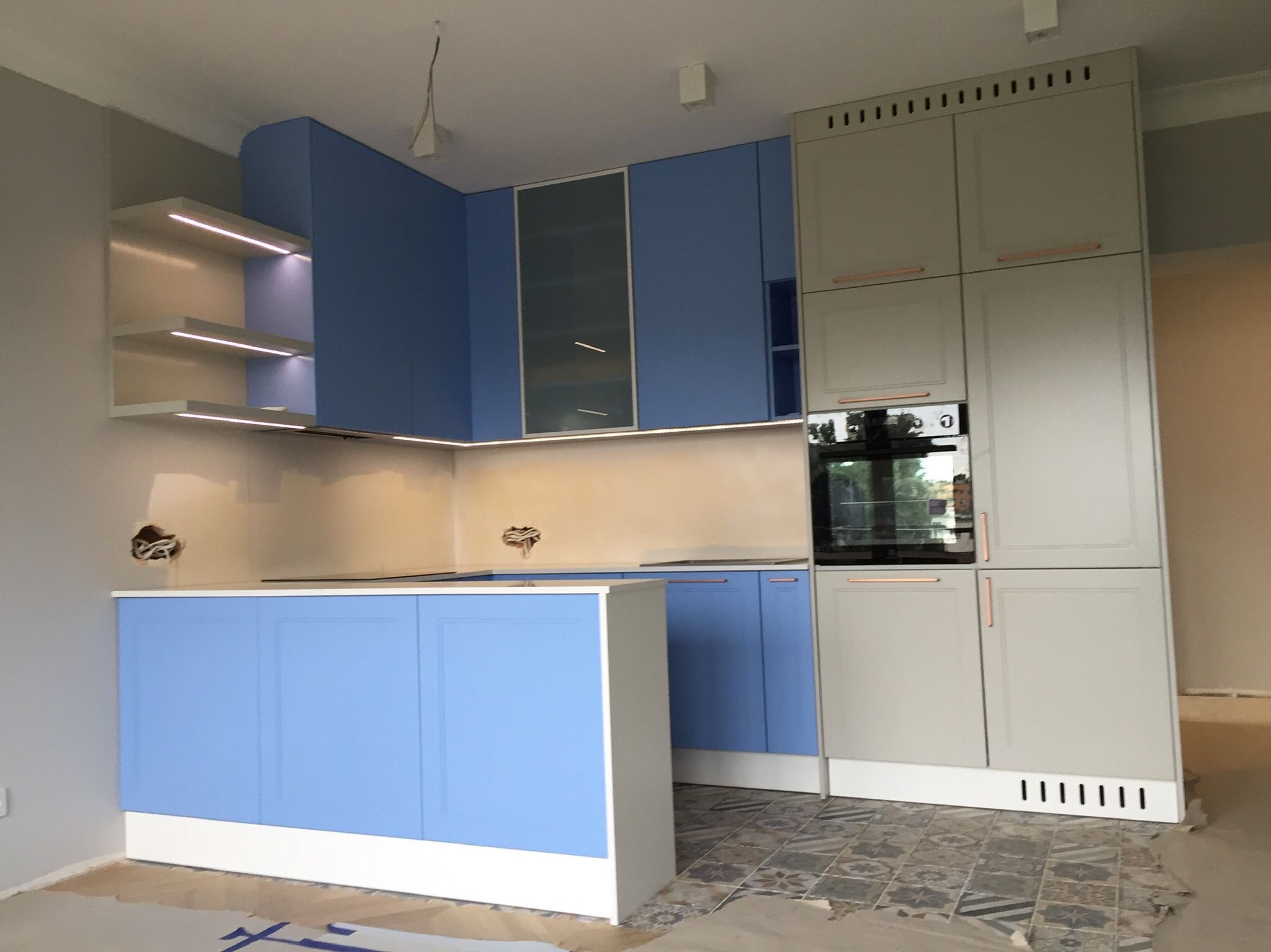 Niebiesko-szara zabudowa kuchenna w trakcie montażu, z oświetleniem LED pod półkami, widoczne niedokończone ściany i zabezpieczona podłoga.