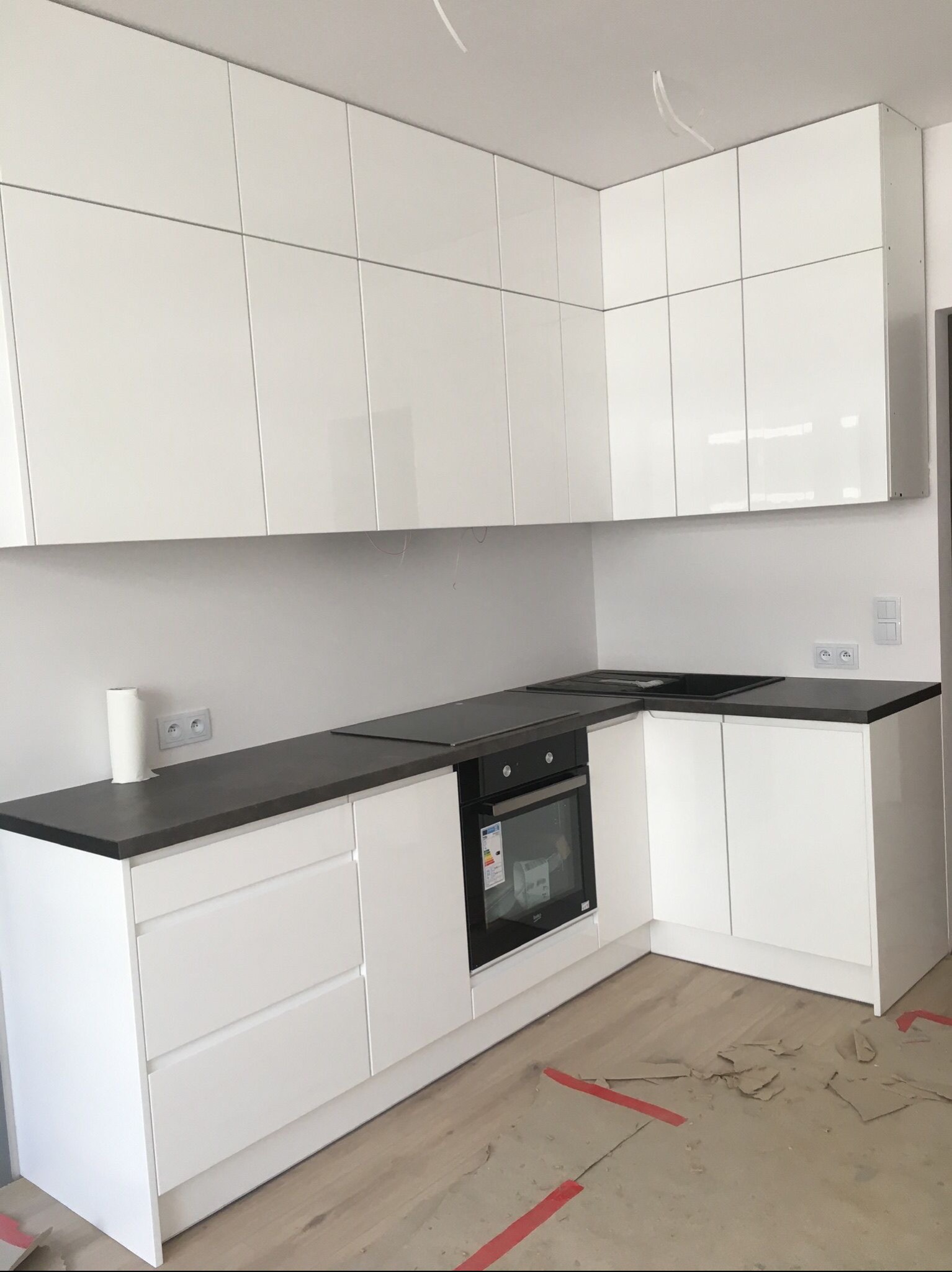 Nowoczesna biała kuchnia z ciemnym blatem, widok na górne i dolne szafki, w tle widoczne kable wystające z sufitu, na podłodze resztki po remoncie.