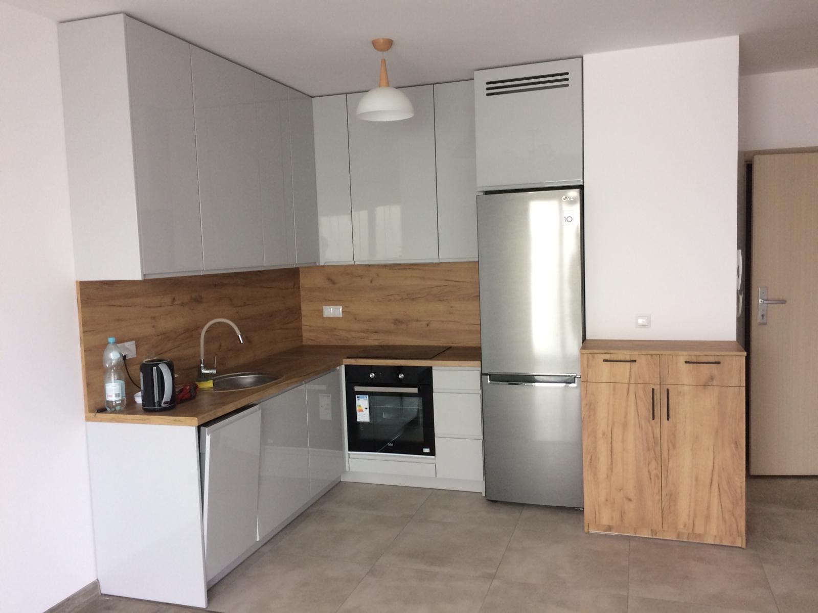 Nowoczesna, jasna kuchnia z szarymi, lakierowanymi frontami szafek, drewnianym blatem i panelem ściennym, srebrną lodówką oraz drewnianą szafką stojącą obok drzwi.