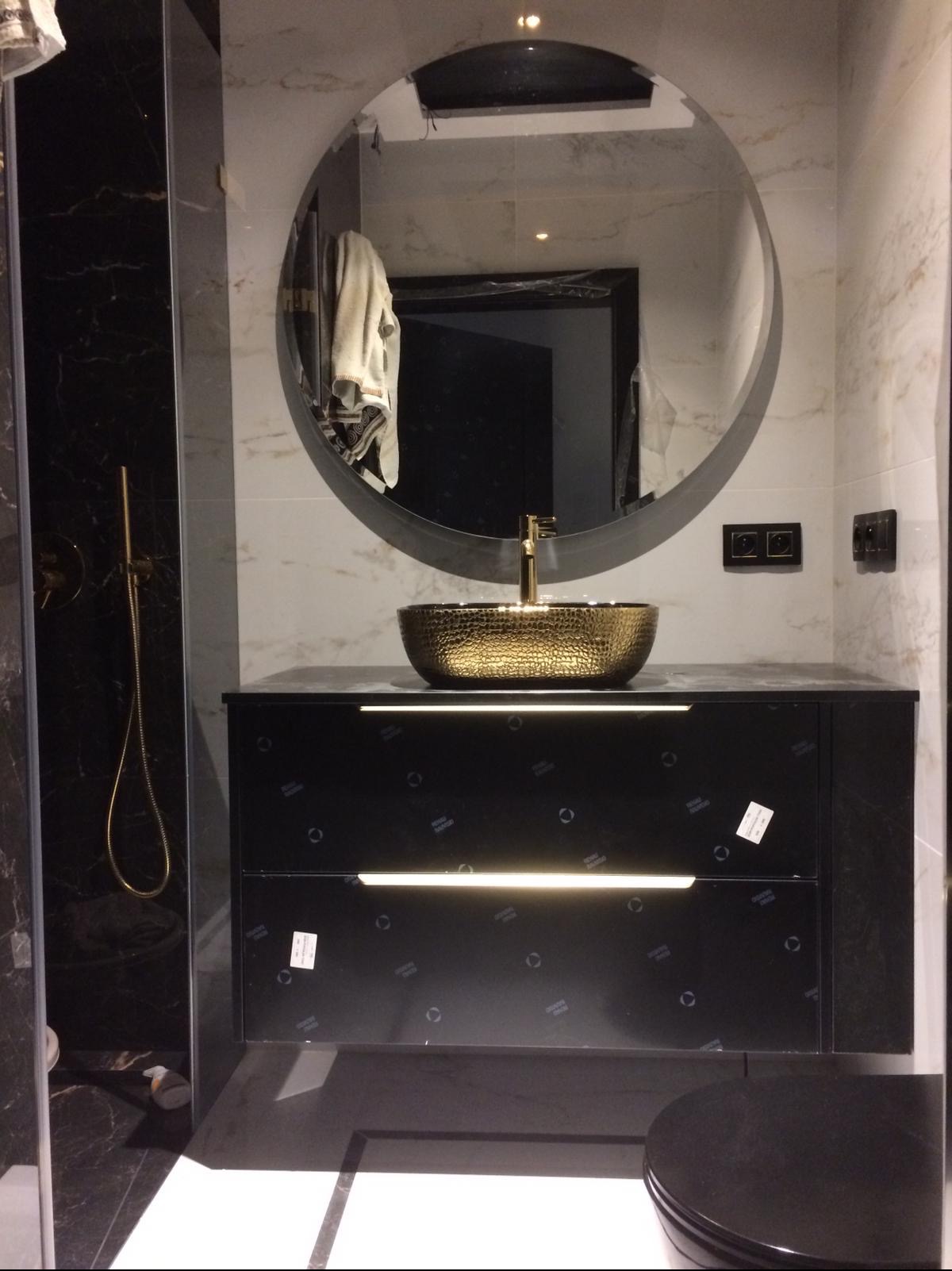 Nowoczesna łazienka z czarną szafką pod umywalkę, złotą, młotkowaną umywalką nablatową i okrągłym lustrem z podświetleniem LED.