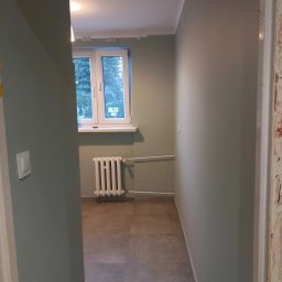 AWANGARDA - Korytarz po odświeżeniu, ściany pomalowane na oliwkowy odcień, widoczny grzejnik pod oknem i szare płytki podłogowe, futryna drzwi w trakcie renowacji.