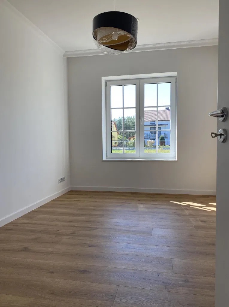 Pusty pokój z drewnianą podłogą i świeżo pomalowanymi ścianami na biało, okno z widokiem na dom sąsiada, lampa wisząca w folii ochronnej, widoczne gniazdka elektryczne.