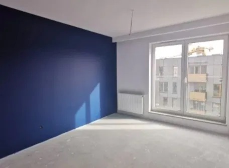 Pomieszczenie po malowaniu: jedna ściana w odcieniu głębokiego granatu, pozostałe ściany białe, duże okno z widokiem na budynek w tle, grzejnik pod oknem, surowa podłoga przed położeniem paneli.