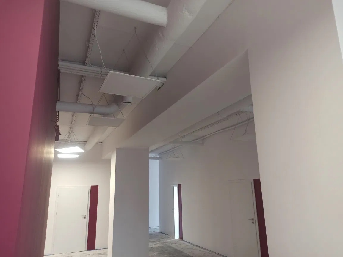 Wnętrze biurowe po remoncie, z pomalowanymi na biało ścianami i sufitem, widoczne instalacje pod sufitem, różowa ściana w tle i drzwi z bordowymi akcentami.