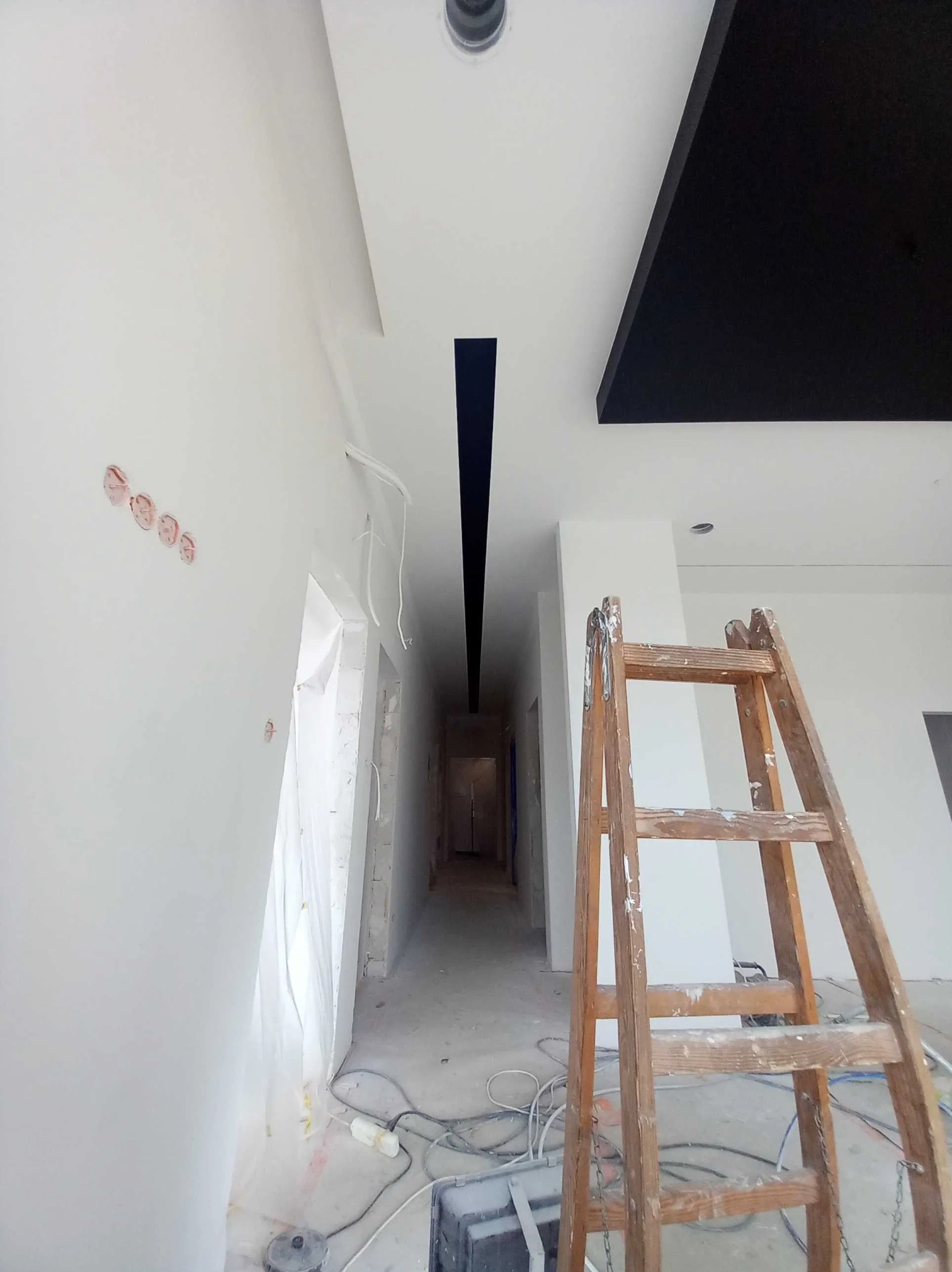 Wnętrze w trakcie prac wykończeniowych: świeżo pomalowana na biało ściana z widocznymi puszkami elektrycznymi, drewniana drabina malarska, fragment czarnego sufitu podwieszanego i długi korytarz...